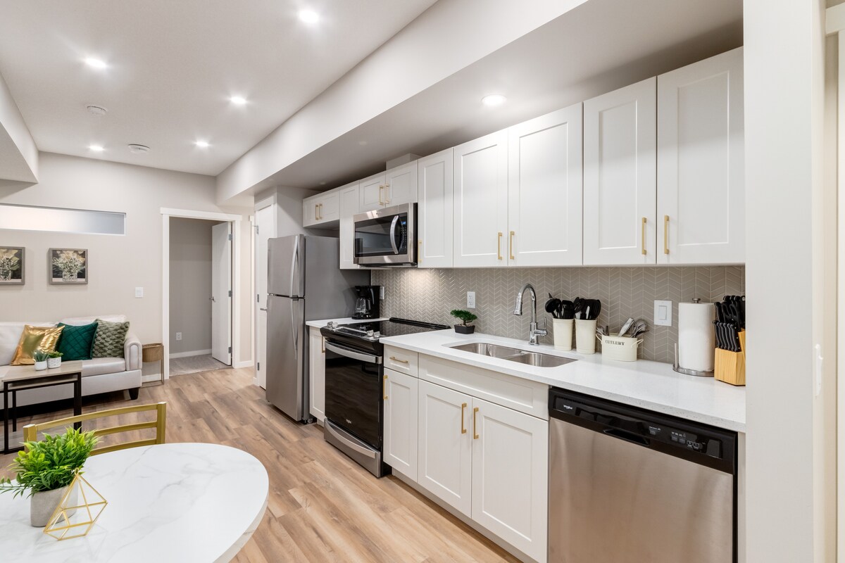 A modern kitchen is displayed with sleek white cabinetry and stainless steel appliances. Ample counter space is provided, along with a dishwasher and a full-sized refrigerator. A stylish dining area with a round table sits nearby, complemented by green and gold decor accents.