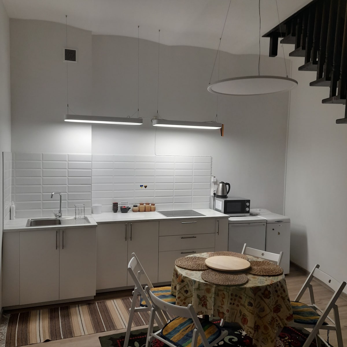 A modern kitchen is equipped with sleek cabinetry and a minimalist countertop. Two pendant lights illuminate a round dining table surrounded by four white chairs. Appliances, including a microwave and refrigerator, complement the functional layout, while a patterned rug adds warmth to the space.