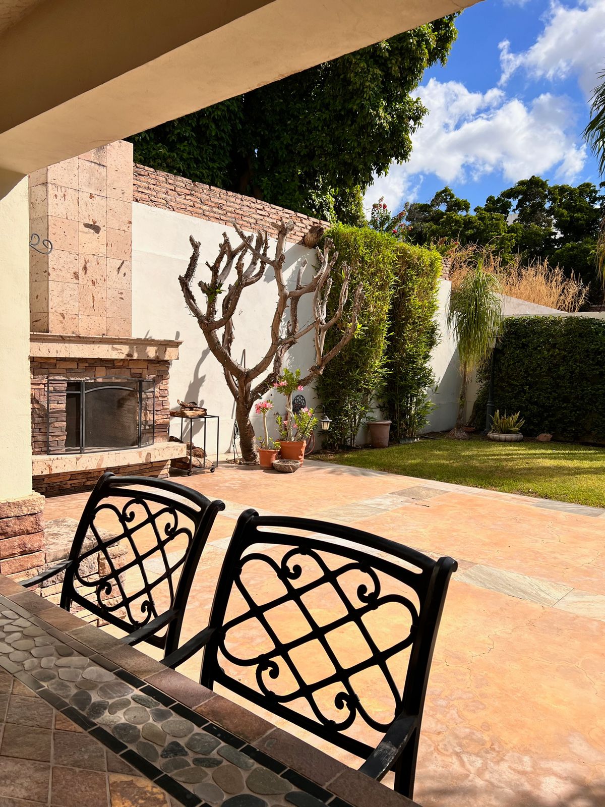 A spacious patio features a stone fireplace and well-maintained greenery. Potted plants are arranged alongside a wall, while a textured stone surface complements the outdoor setting. Two iron chairs circle a table, providing a comfortable area for gathering.