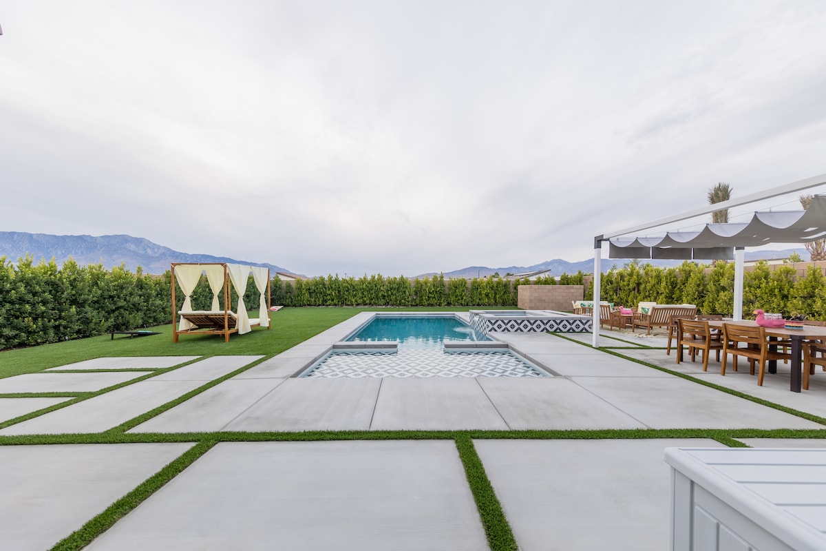 The outdoor area features a rectangular pool surrounded by a smooth patio with geometric patterning. A dining area is visible, equipped with a wooden table and chairs. Nearby, a shaded lounging space offers comfortable seating, while lush greenery creates a private atmosphere with mountain views in the background.