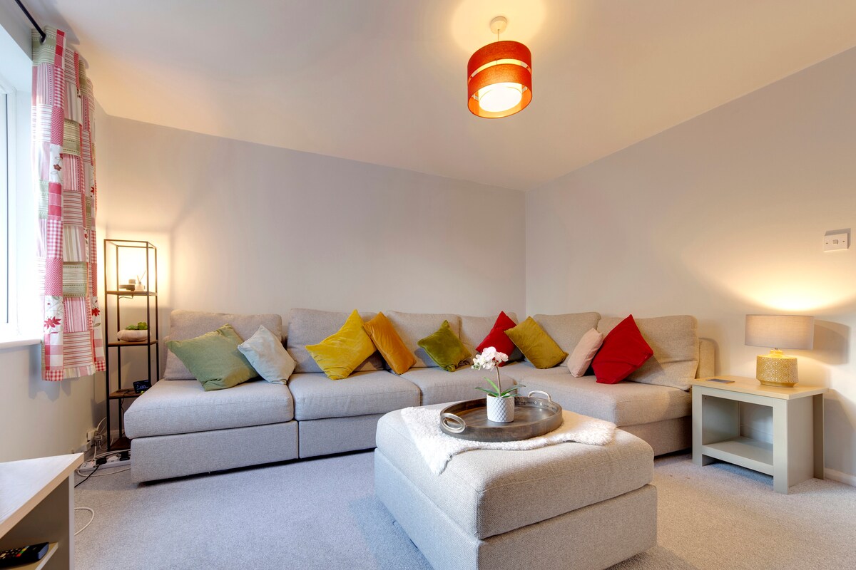A light-filled living room features a spacious sectional sofa adorned with colorful cushions. A round coffee table is accented with a small plant and tray. Soft gray walls and a warm overhead light create an inviting space. A floor lamp adds additional illumination.