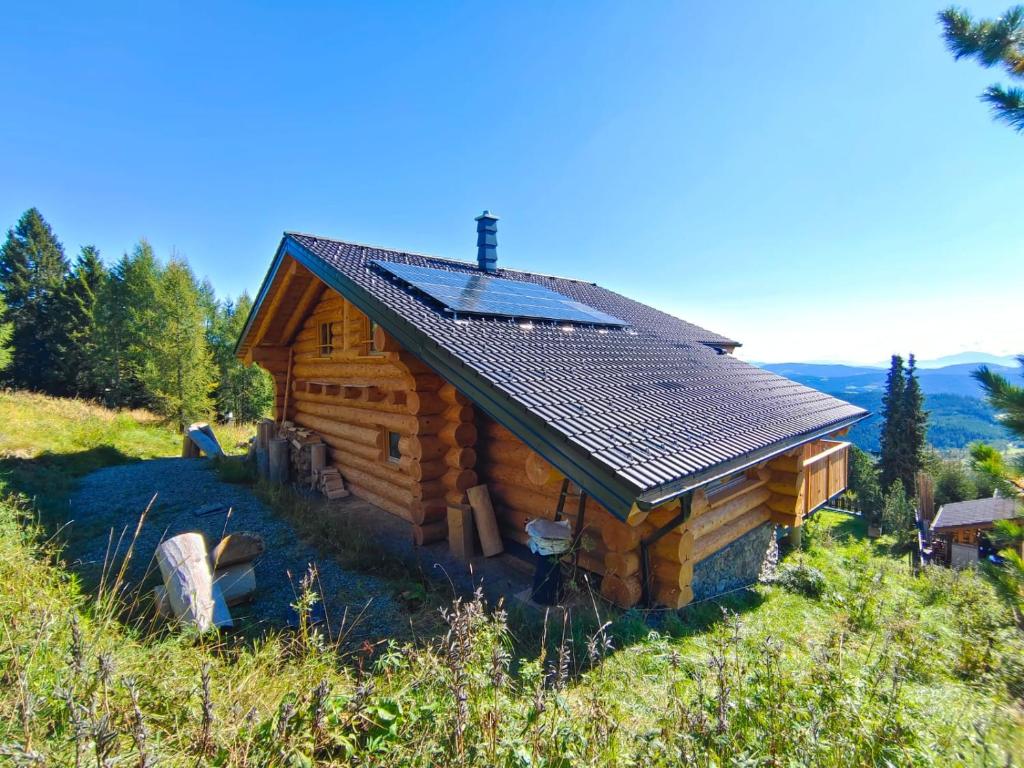 A rustic log cabin is nestled amidst greenery, showcasing a sloped roof with solar panels. The facade features wooden logs and natural stone accents, surrounded by tall trees and open landscapes, while outdoor seating options are situated nearby.