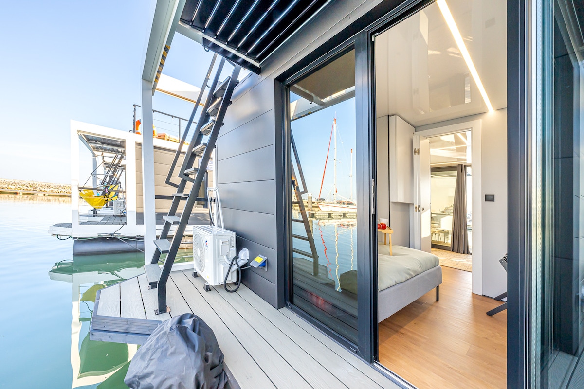 The image captures a modern homeboat with a spacious deck leading to large glass doors. Inside, a well-lit sleeping area is visible, featuring a comfortable bed. The serene water reflects the clear blue sky, and the marina is seen in the background.