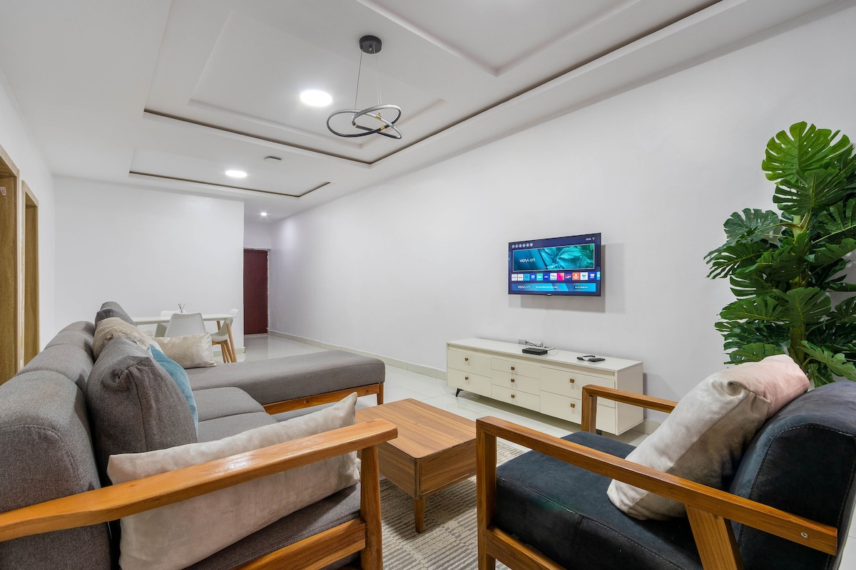 A spacious living area is presented with a combination of grey and black sofas, complemented by a wooden coffee table. A flat-screen television is mounted on a light wall, while a large green plant adds a touch of nature to the ambiance.
