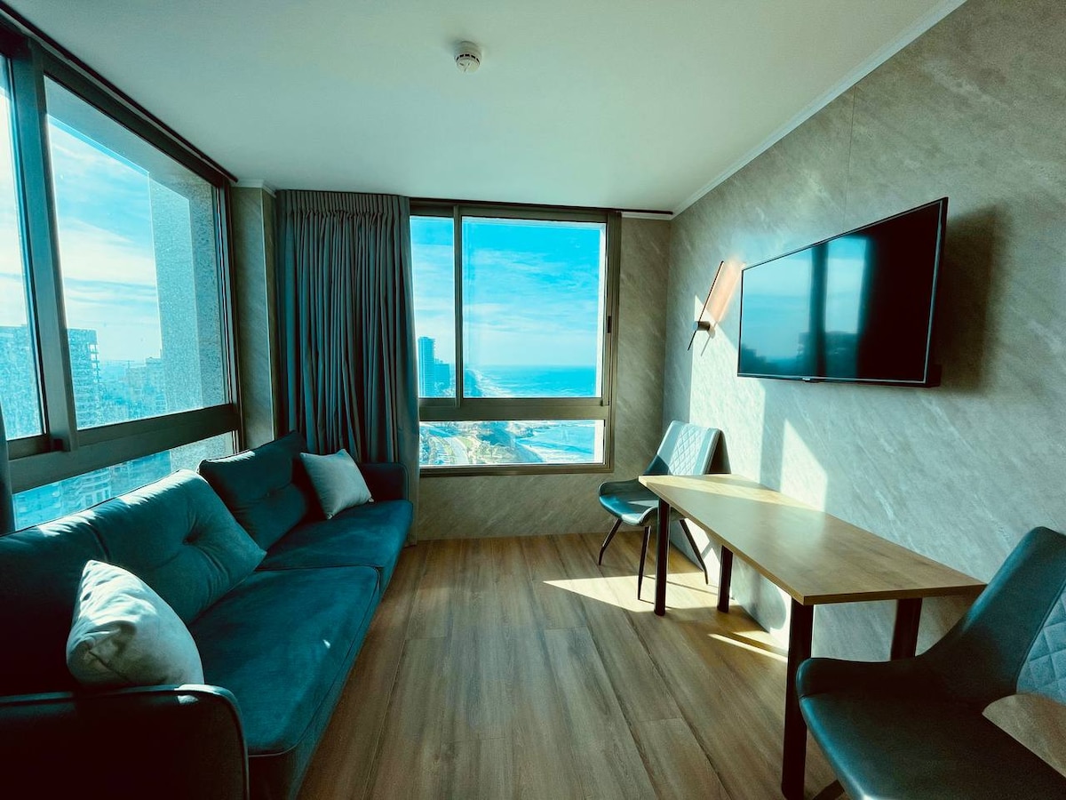 A light-filled living space features large windows offering views of the ocean. A cozy sofa is positioned beside a wooden table, while two chairs complement the seating area. A modern television is mounted on the wall, enhancing the open ambiance.