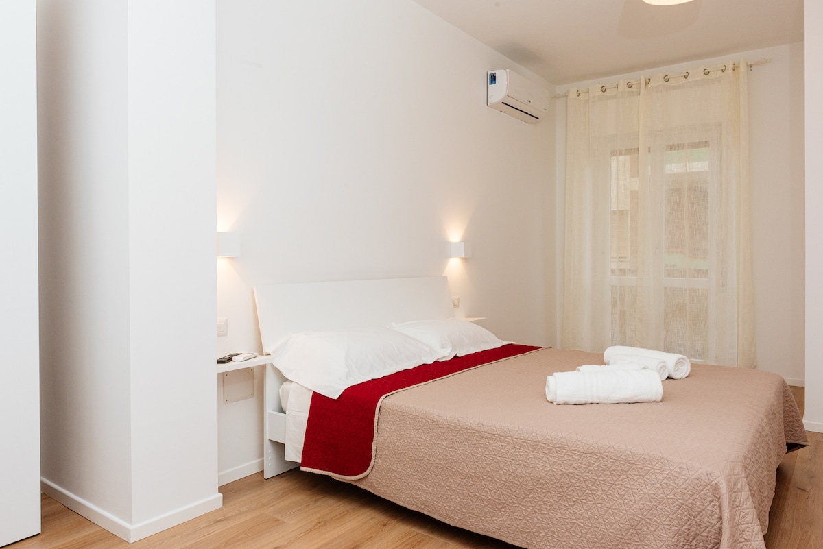 A bright bedroom is highlighted by soft, neutral walls and warm wooden flooring. A spacious bed is dressed in a light quilt with a contrasting red throw, complemented by neatly folded towels. A large window is adorned with sheer curtains, allowing natural light to fill the room.
