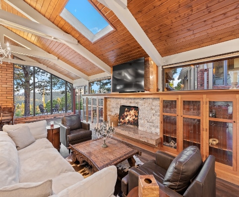 A spacious living area features high ceilings with wooden beams and skylights allowing natural light. A stone fireplace serves as the focal point, complemented by comfortable seating arrangements. Large windows provide panoramic views of the surrounding landscape.