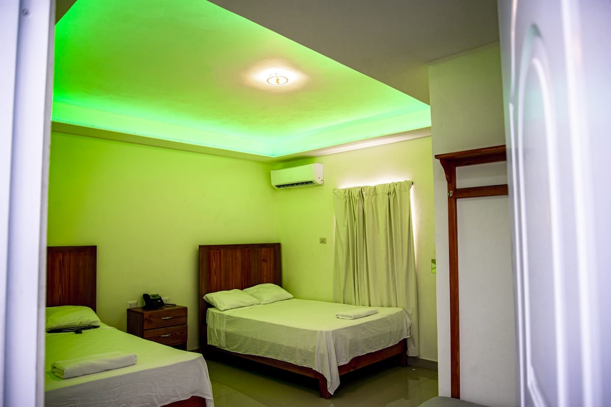 A well-lit room features two spacious beds with simple linens, set against light green accent lighting above. A wooden nightstand and a small phone are visible beside one bed. Flowing curtains partially cover a window, complementing the room's calming atmosphere.