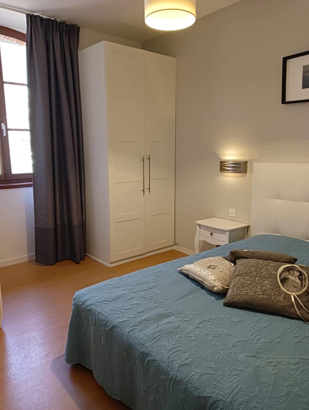 A well-organized bedroom features a white bed with a textured blue coverlet and decorative pillows. A bedside table and a wardrobe provide storage. Natural light enters through the window, which is dressed with light curtains, adding a soft ambiance to the space.