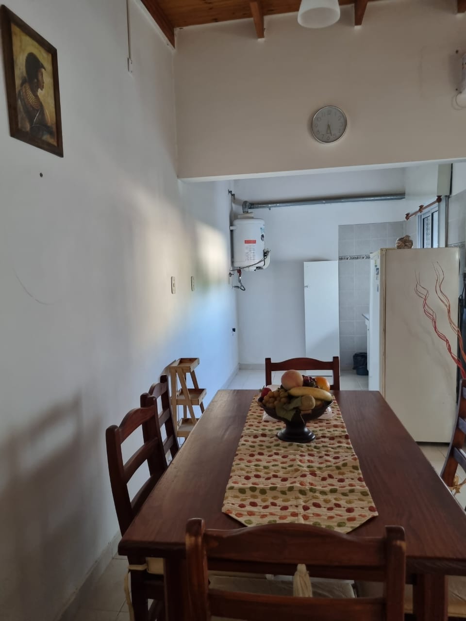 A wooden dining table is set with a decorative bowl of fruit, surrounded by chairs. A patterned table runner extends across the table. The room features white walls, a clock mounted on the wall, and a glimpse of the kitchen area in the background.