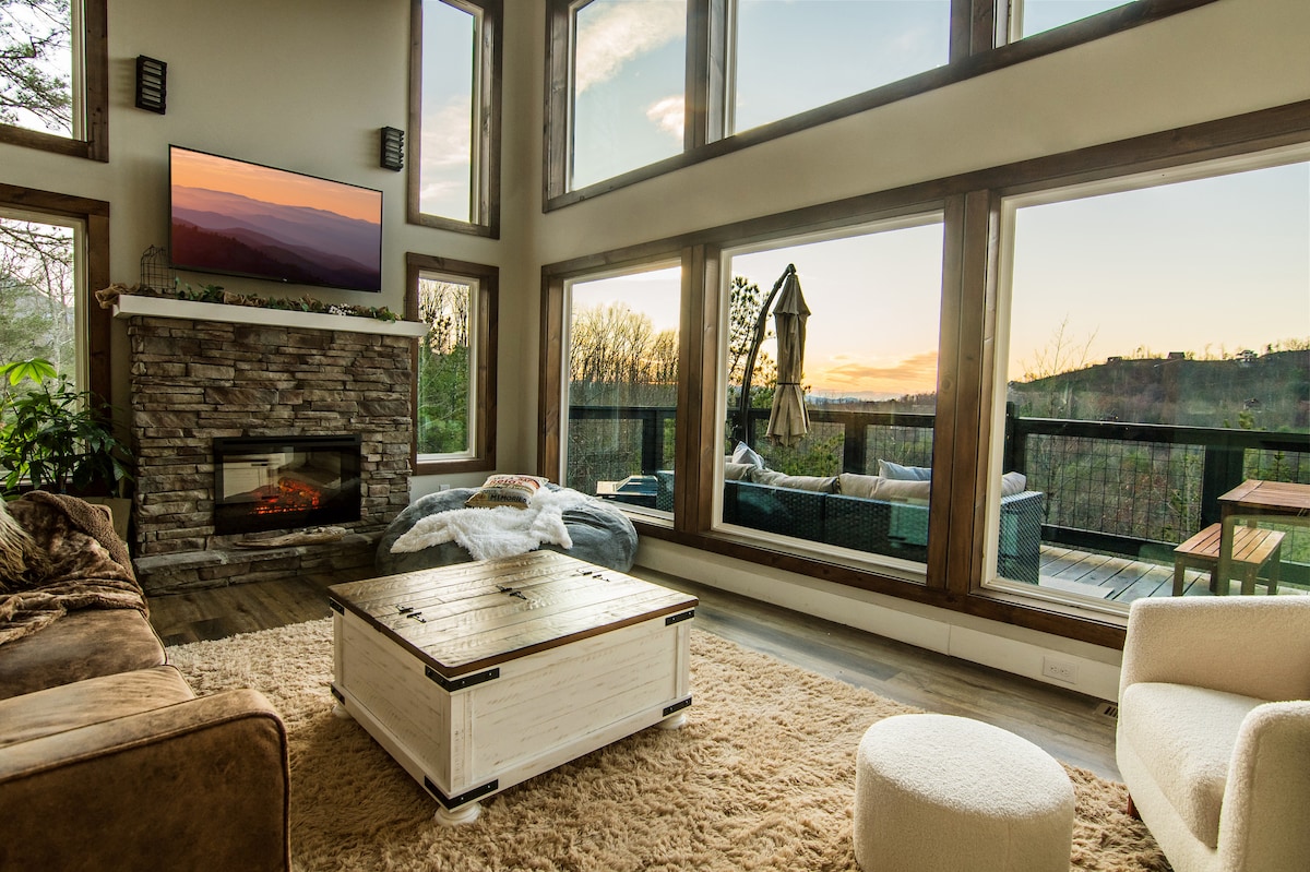 A spacious living area features large windows offering mountain views at sunset. A mounted TV is above a stone fireplace, and a cozy sectional sofa is paired with a rustic coffee table. A soft area rug enhances the warmth of the room.