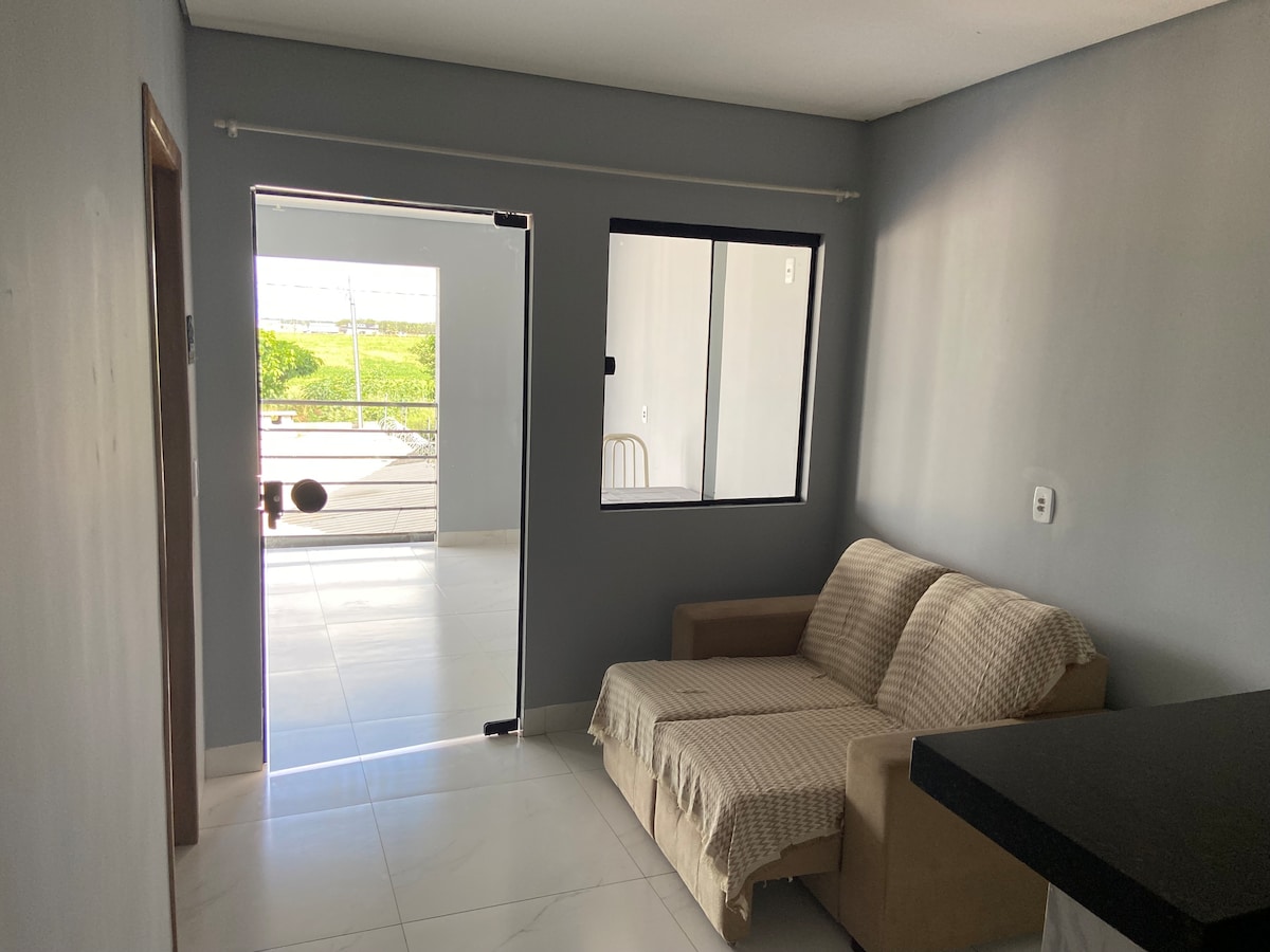 A cozy living area is depicted, featuring a beige sofa draped with a light blanket. An adjacent glass door invites natural light, leading to a balcony with views of greenery. Gray walls and glossy white tiles enhance the open space's modern feel.