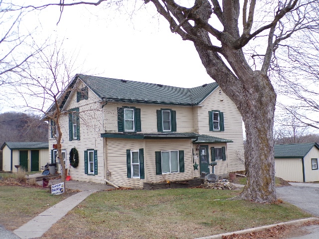 A two-story house features a light-colored exterior with dark green shutters. A large tree sits in the foreground, adding shade to the front yard. The structure is complemented by a stone pathway leading to the entrance, showcasing the inviting layout of the property.