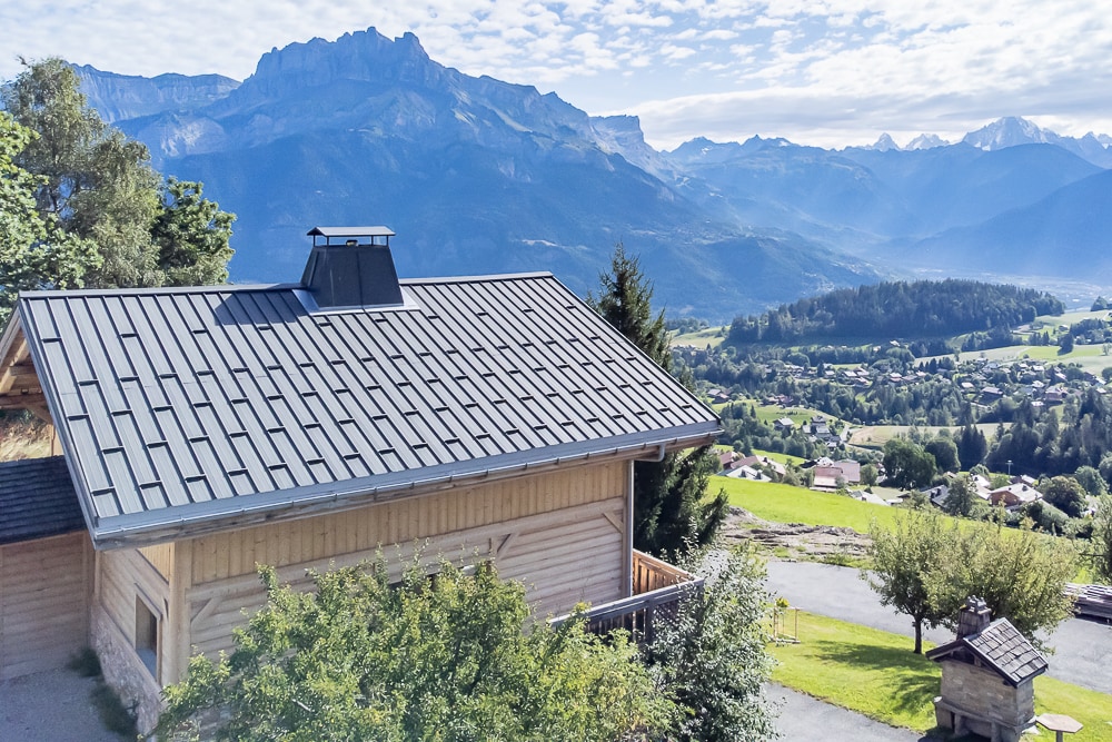 The exterior of the chalet is captured against a backdrop of majestic mountains and valleys. The wooden construction features a modern roof with a chimney. Lush greenery surrounds the property, and panoramic views of the landscape are visible in the distance.