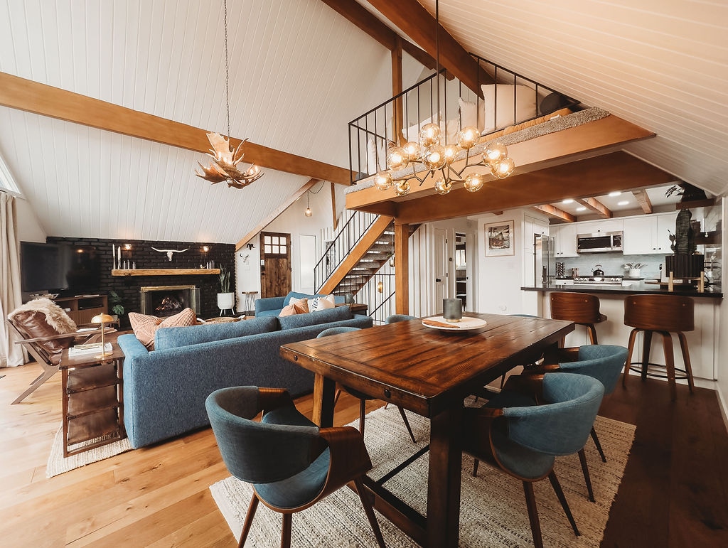 A spacious living and dining area features a large wooden dining table surrounded by blue upholstered chairs. A cozy blue sofa is arranged with leather chairs, providing a warm gathering space. High ceilings and large windows enhance the bright ambiance, while a modern chandelier adds style.