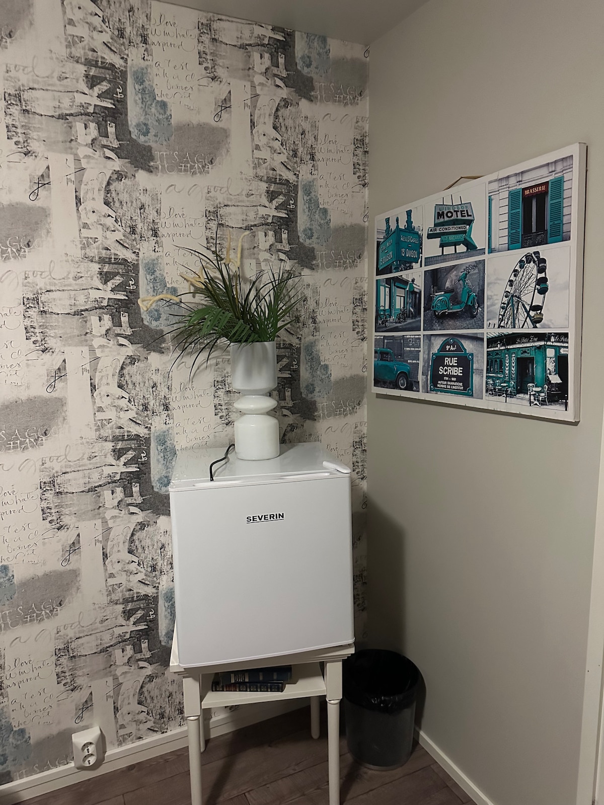 A corner of the room features a small white refrigerator on a slender stand, adorned with a green plant in a vase. A wall adorned with patterned wallpaper complements a framed collage of vintage photographs, including a ferris wheel and a retro motel.