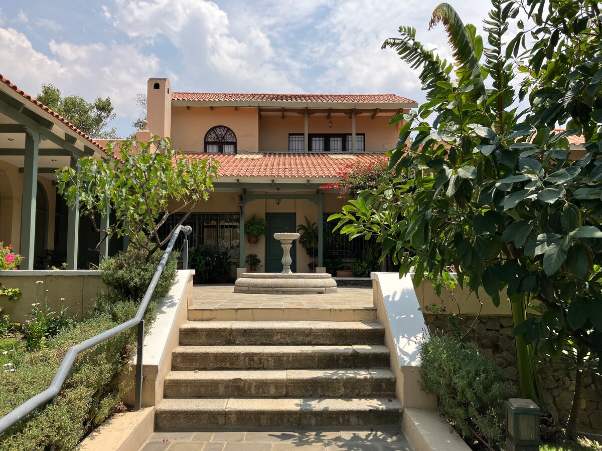 A welcoming entrance features a set of stone steps leading to a charming house with a red-tiled roof. Lush greenery surrounds the space, including trees and colorful plants, while a central fountain adds a touch of elegance to this serene outdoor area.