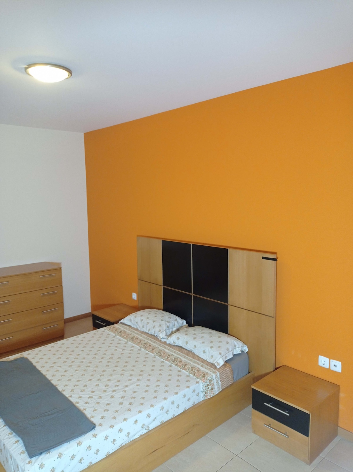 A cozy sleeping area features a spacious bed with a patterned duvet and two nightstands. An orange accent wall adds a pop of color, while a wooden dresser is positioned against the opposite wall, providing additional storage.