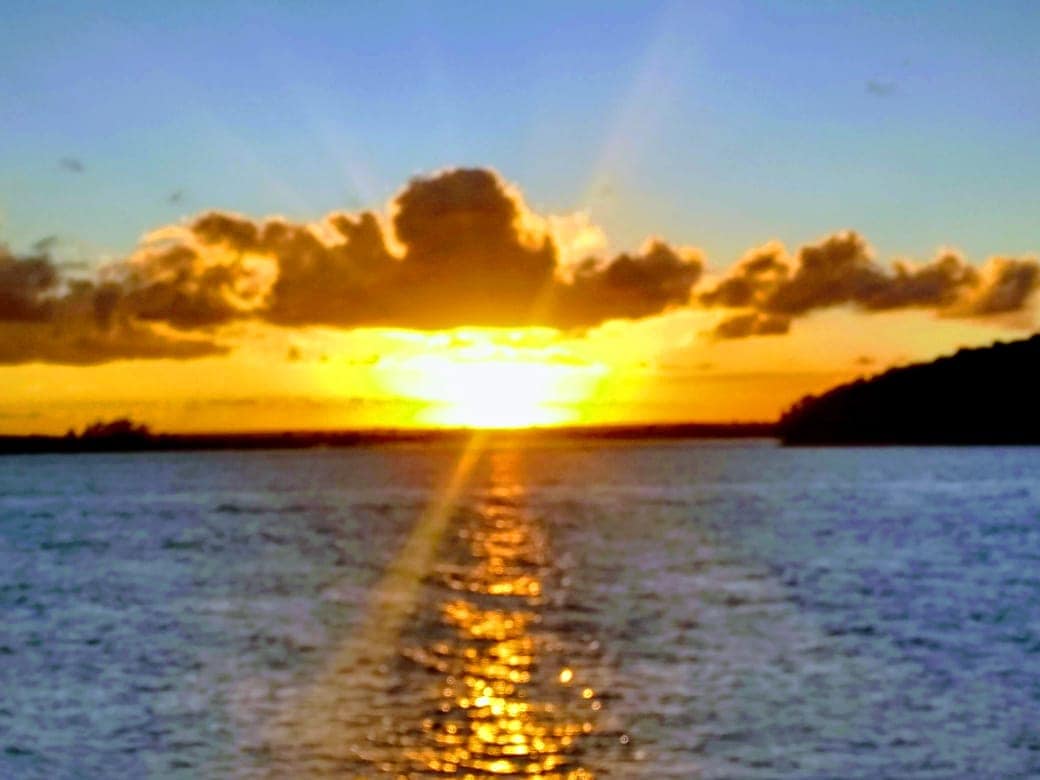 A serene sunset is captured over calm waters, with the sun dipping below the horizon. Soft orange and yellow hues emanate from the sun, reflecting on the water's surface. Fluffy clouds are visible, creating a tranquil backdrop against a peaceful sky.