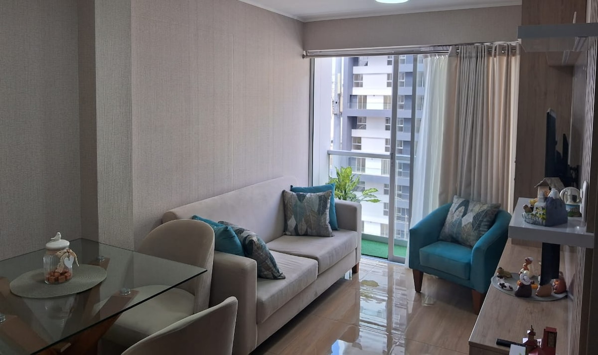 A well-lit living area features a light-colored sofa adorned with decorative pillows. A glass dining table is positioned alongside comfortable chairs. A vibrant blue armchair adds a pop of color. Large windows allow natural light to illuminate the space, with a view of greenery outside.
