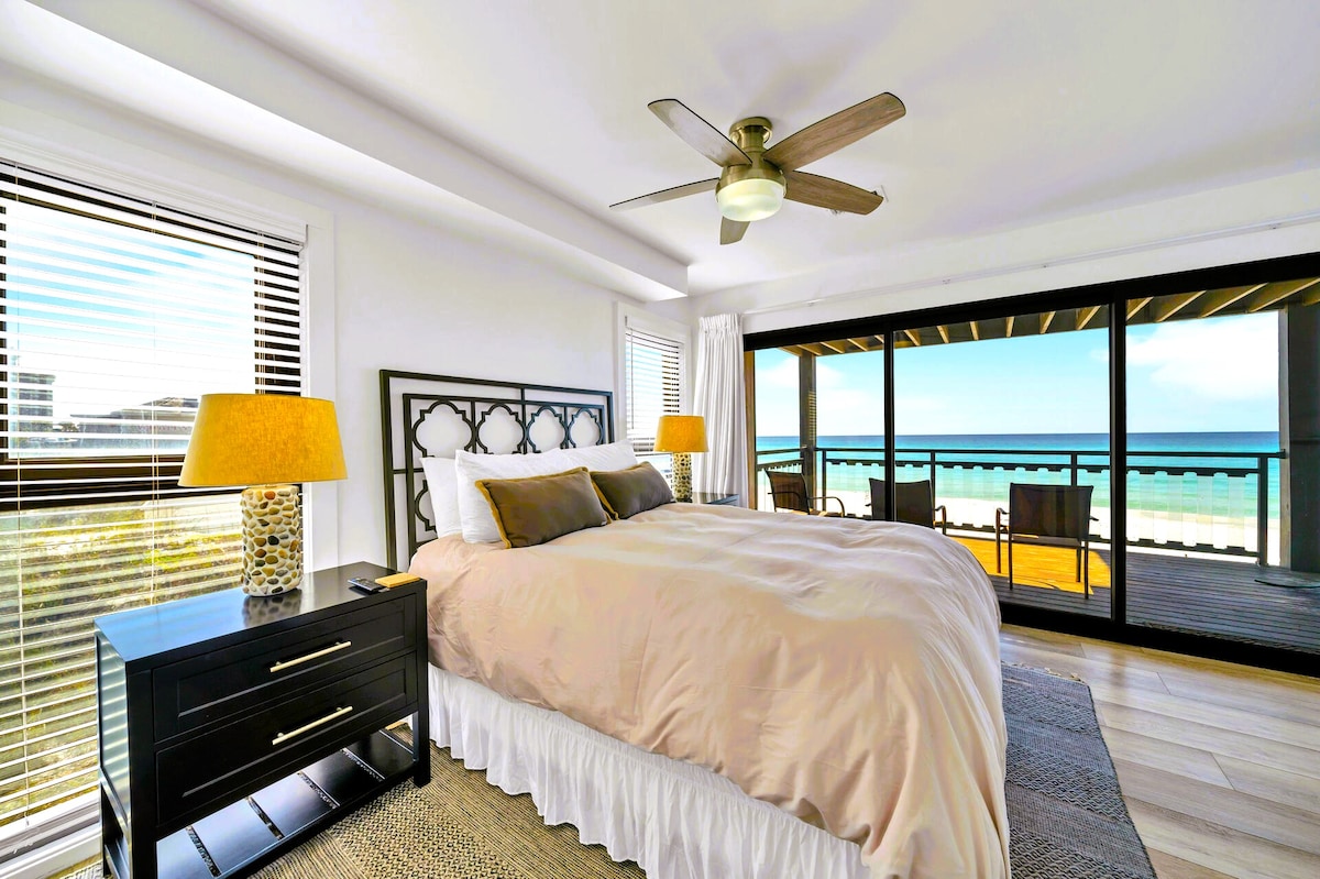 A spacious bedroom features a large bed adorned with neutral bedding. A bedside table with a lamp provides soft lighting. Large windows offer views of the ocean, while a private balcony is visible, enhancing the serene atmosphere.