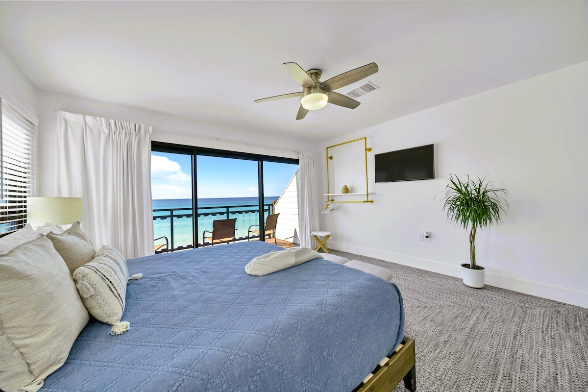 A spacious bedroom features a comfortable bed with a blue quilt and decorative pillows. Large glass doors provide an unobstructed view of the ocean. A ceiling fan circulates air, and a potted plant adds a touch of greenery. A small flat-screen TV is mounted on the wall.