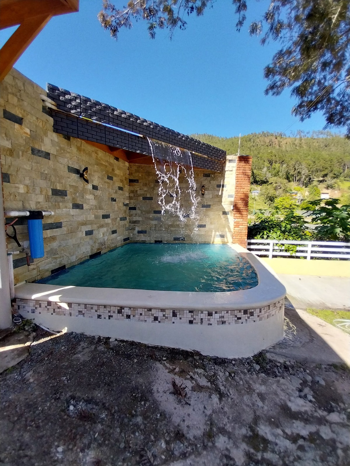 A private outdoor pool is surrounded by natural stone walls and accented by cascading water features. Sunlight reflects off the clear water, inviting relaxation in the serene setting of the villa's garden.