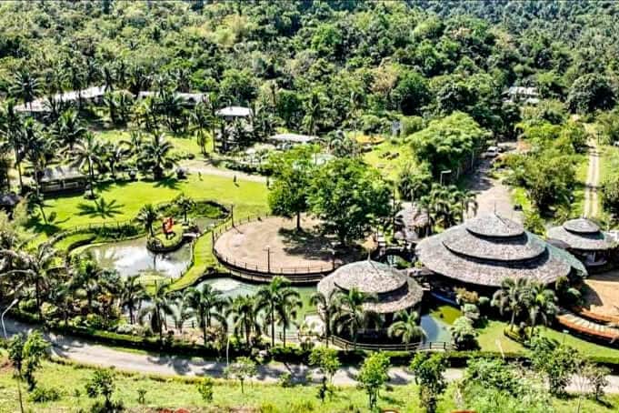 An aerial view showcases the resort's layout, featuring several thatched-roof structures surrounded by lush greenery. A serene pond reflects the landscape, while pathways meander throughout the expansive grounds, lined with palm trees and vibrant landscaping.