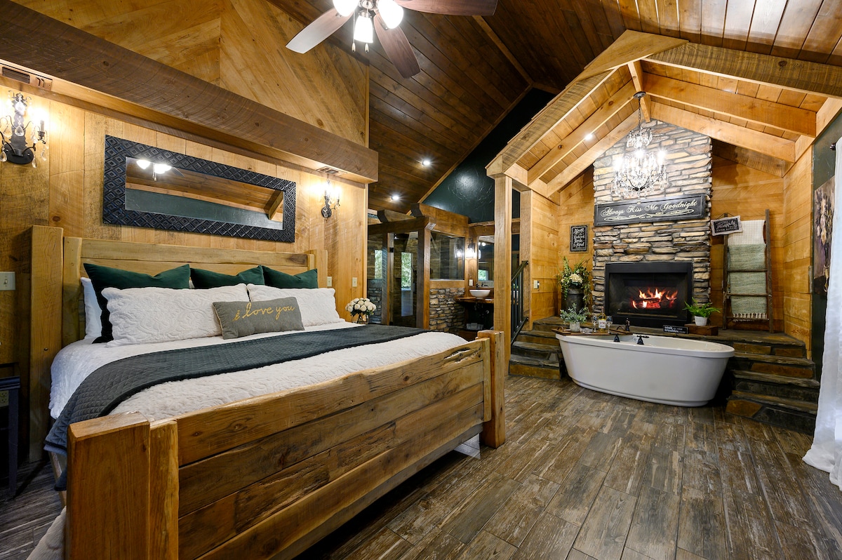 A spacious bedroom features a wooden bed framed with decorative pillows and a textured blanket. A rock fireplace is positioned next to a sleek, freestanding bathtub. Natural wood accents and a high ceiling enhance the rustic charm, while soft lighting creates a cozy ambiance.
