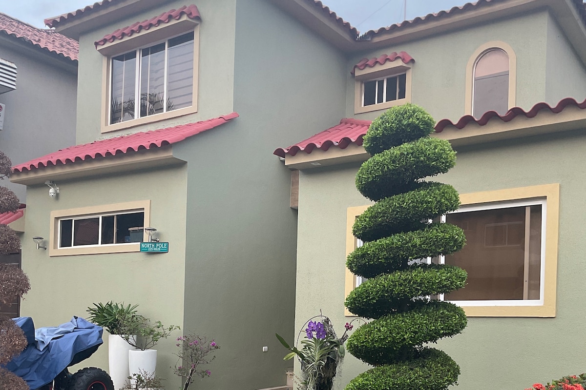 The exterior of the villa is showcased, highlighting its two-story design with a tiled roof and a mix of green and beige walls. A manicured topiary stands in the foreground, complemented by blooming plants and a parked vehicle covered by a blue tarp.