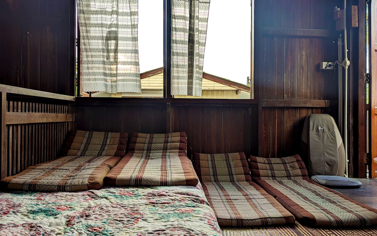 An inviting sleeping area is equipped with several checkered floor cushions arranged on woven mats. Light filters through patterned curtains, illuminating the rustic wooden walls. A soft quilt adds texture to the space, and a few personal items are neatly placed in the corner.