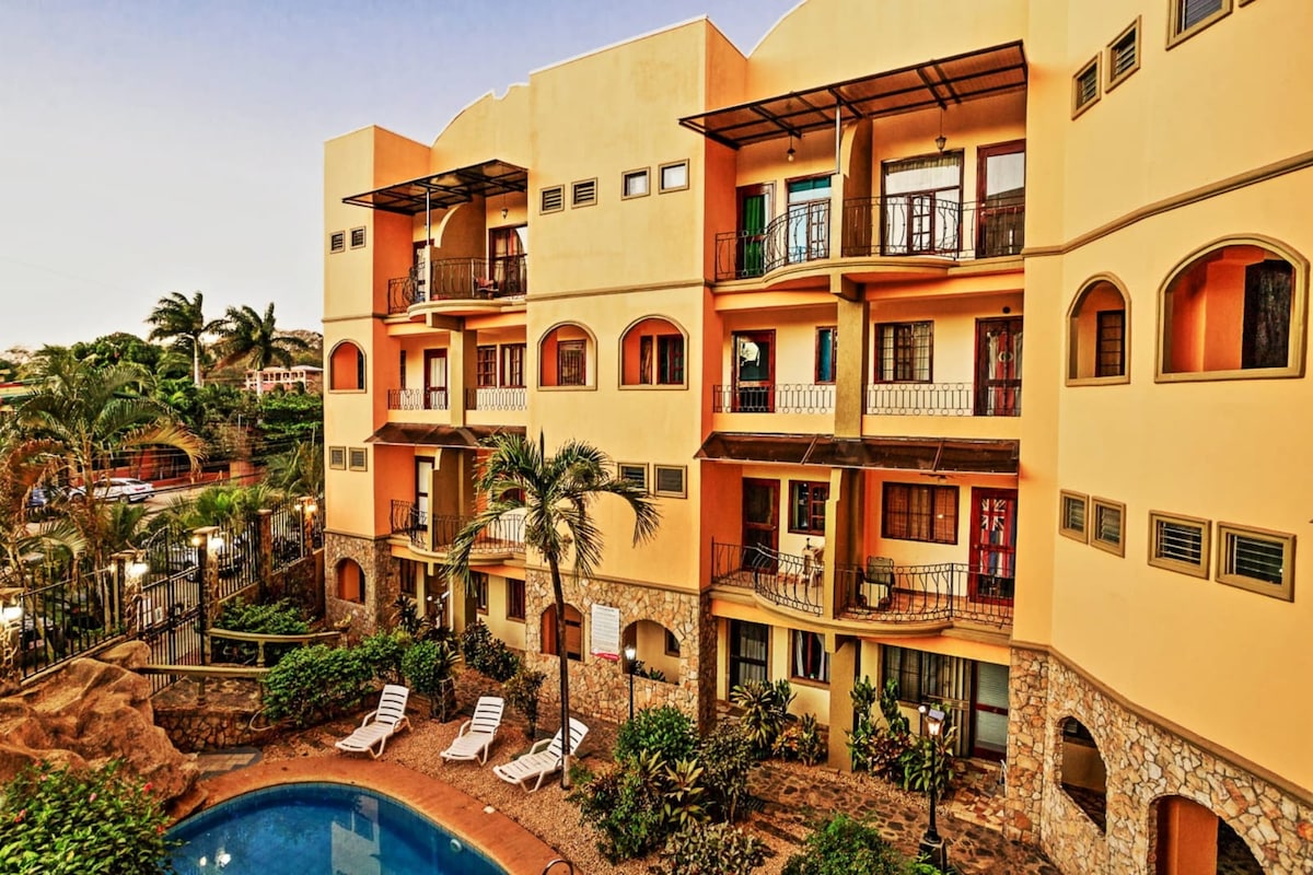 The image showcases a vibrant, two-story condo building painted in warm tones, surrounded by lush greenery. Balconies adorn each unit, offering views of the central pool area. Sun loungers are positioned near the inviting pool, creating a tranquil ambiance in the outdoor space.