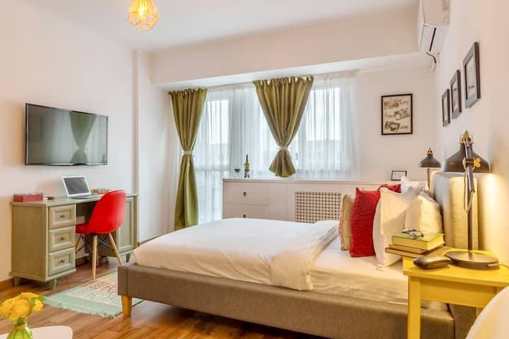 Elegantná spálňa spájajúca pohodlie a funkčnosť s mäkkou manželskou posteľou a vankúšmi. Ponúka elegantný stôl s namontovaným televízorom, príjemné osvetlenie pri posteli a štýlový dekor. Veľké okno so závesmi zapĺňa miestnosť svetlom.