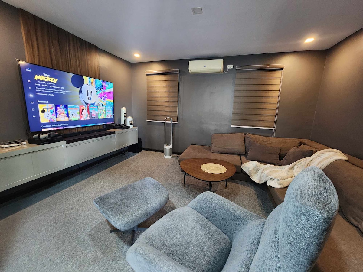A comfortable seating area is featured with a large sofa, an armchair, and a round coffee table. A flat-screen television displays colorful cartoons on the wall, while two windows with shades allow soft lighting into the room.
