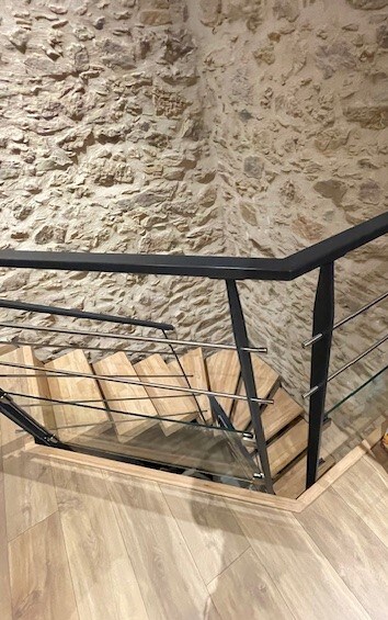 A staircase is visible, featuring wooden steps and a contemporary black railing. The walls exhibit exposed stone, providing texture, while the floor is finished in light wood, enhancing the natural ambiance of the interior.