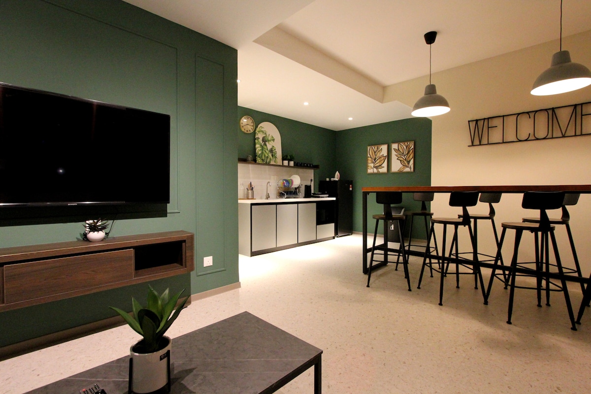 A modern living area features a dark green accent wall, complemented by a sleek entertainment unit with a large TV. The kitchen area includes a minimalistic design with essential appliances visible. A dining table with high chairs and decorative wall art creates a welcoming ambiance.