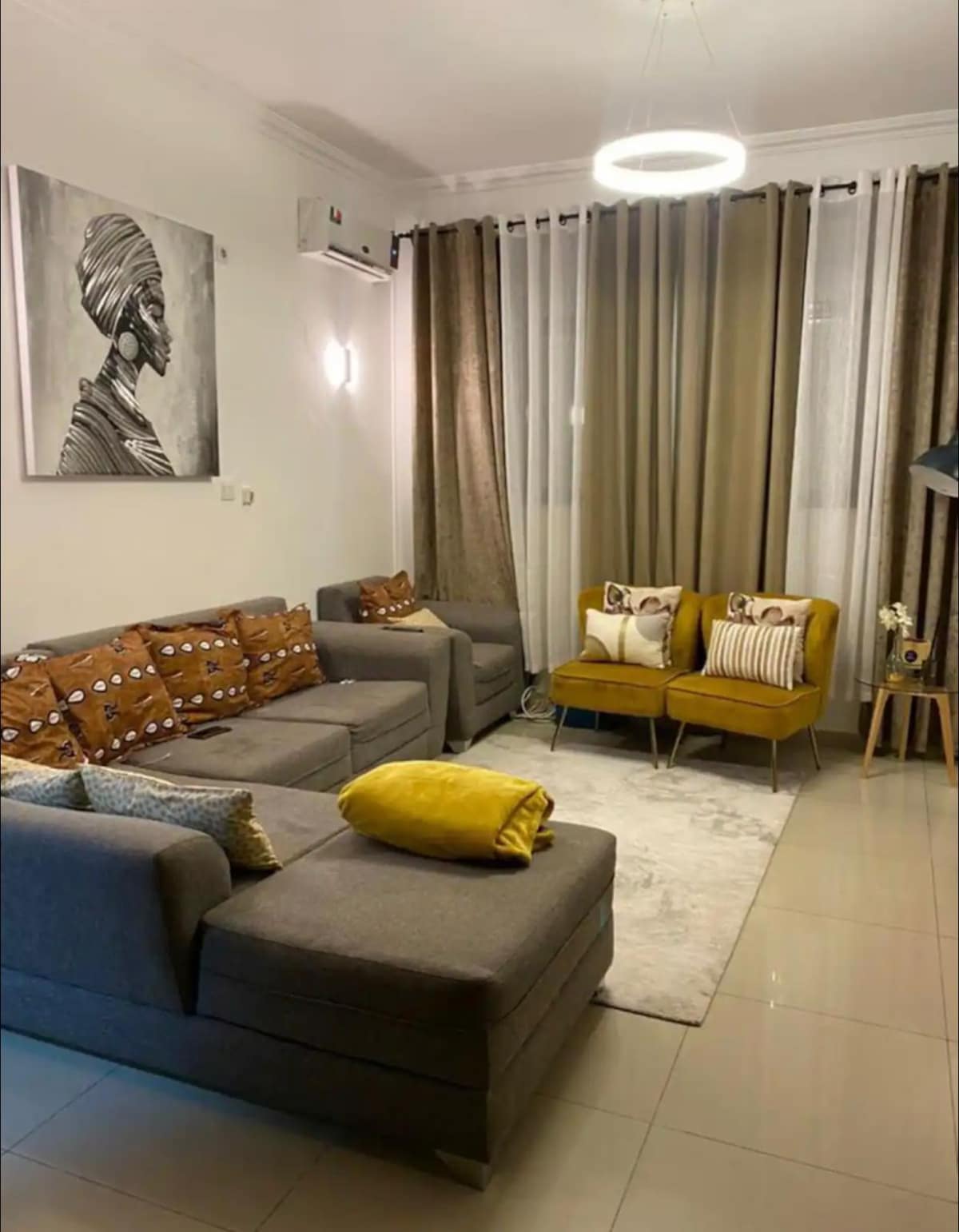 A modern living area features a sectional sofa adorned with patterned cushions and a cozy throw. Light-colored curtains drape around a large window, and two yellow accent chairs provide additional seating. A decorative area rug adds warmth to the tiled floor.