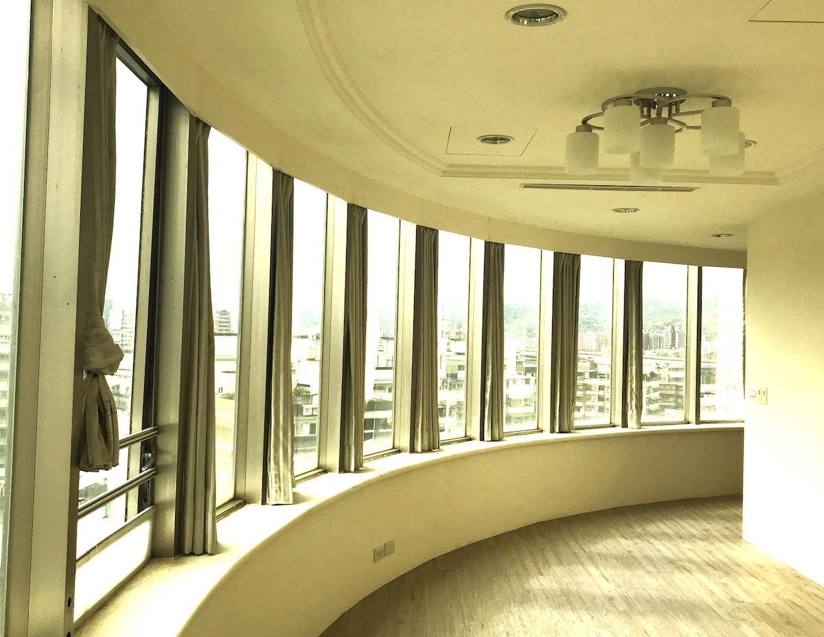 Large windows curve around the space, offering views of the cityscape. Sheer curtains allow natural light to filter through, creating a bright atmosphere. The smooth wooden floor complements the modern design, enhancing the openness of the room.