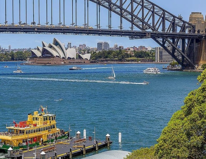 A panoramic view captures the Sydney Harbour featuring the iconic Harbour Bridge and the Sydney Opera House. Calm waters reflect the surroundings, with boats sailing peacefully. A vibrant ferry is docked at a nearby marina, framed by lush greenery along the shoreline.