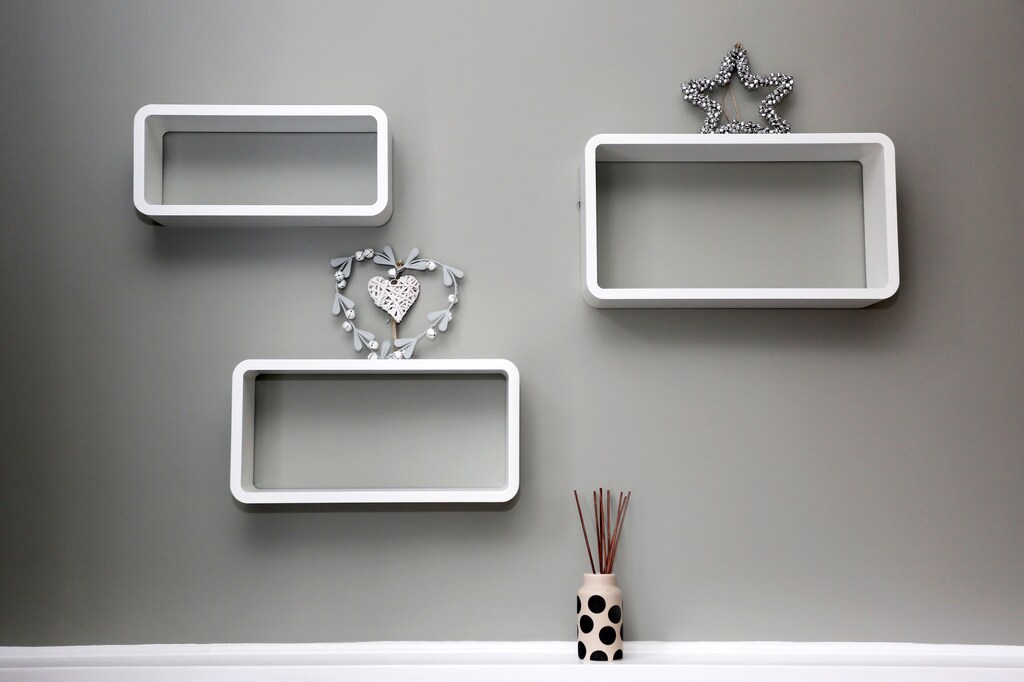 Three white wall-mounted shelves are displayed on a soft gray wall. One shelf features a decorative heart and another has a star accent. A black and white patterned vase containing reed sticks sits below, adding a touch of texture to the arrangement.
