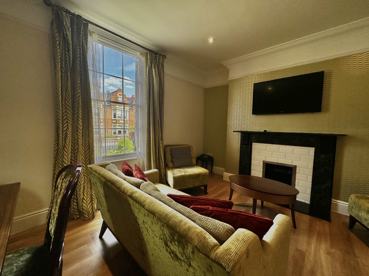 A modern living room features a plush sofa in soft green hues, paired with an inviting round coffee table. Natural light filters through large windows, adorned with elegant drapes. A flat-screen television is mounted above a sleek fireplace, creating a cozy focal point in the room.