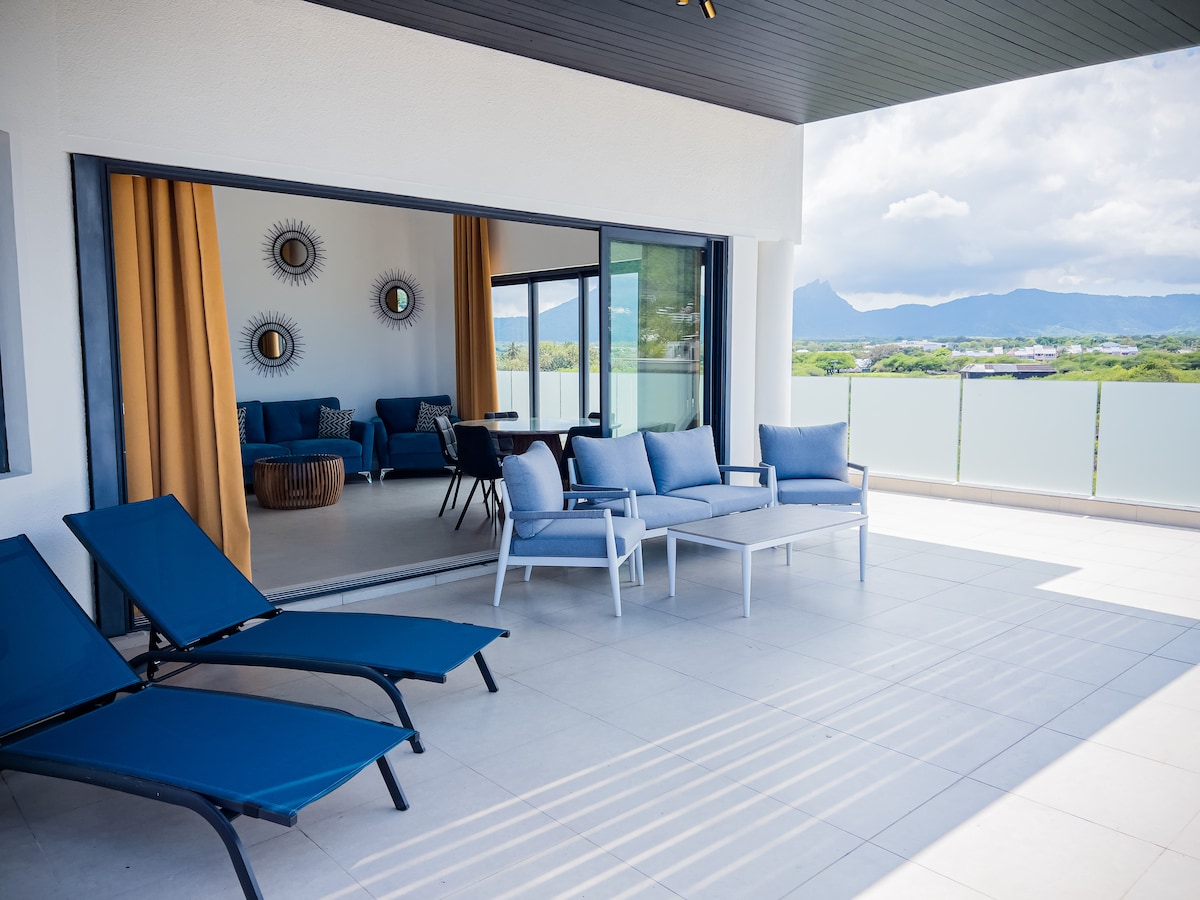 A spacious terrace is presented with modern outdoor furniture, including blue lounge chairs and a table set. Large glass doors provide an unobstructed view of the surroundings. The area features a clean, minimalistic design with neutral-colored walls and accent mirrors to enhance the ambiance.