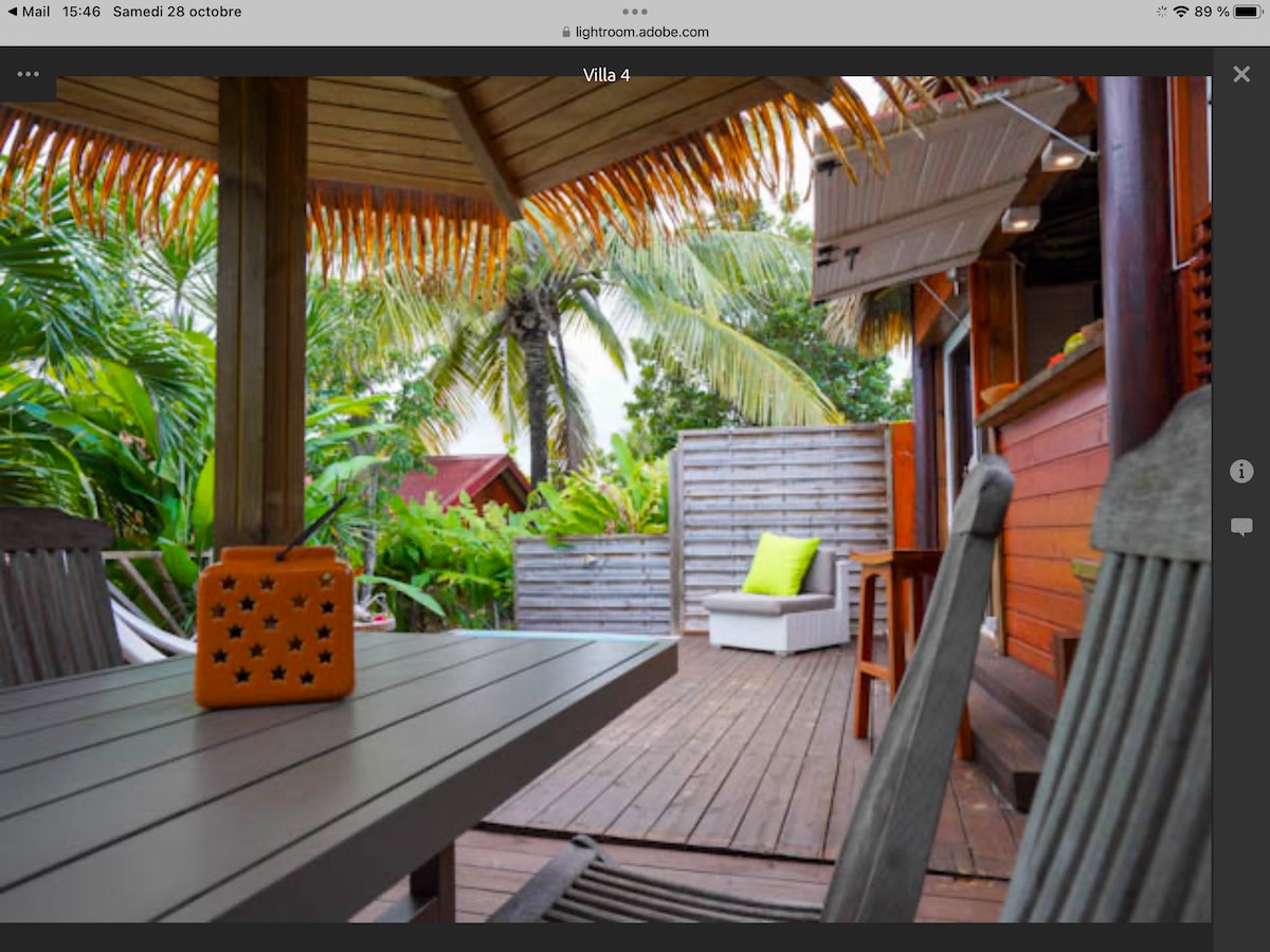 A wooden deck features a dining table set for outdoor meals, accompanied by gray wooden chairs. A brightly colored lantern stands on the table. In the background, a lounge chair is situated against a backdrop of lush greenery and tropical palms.