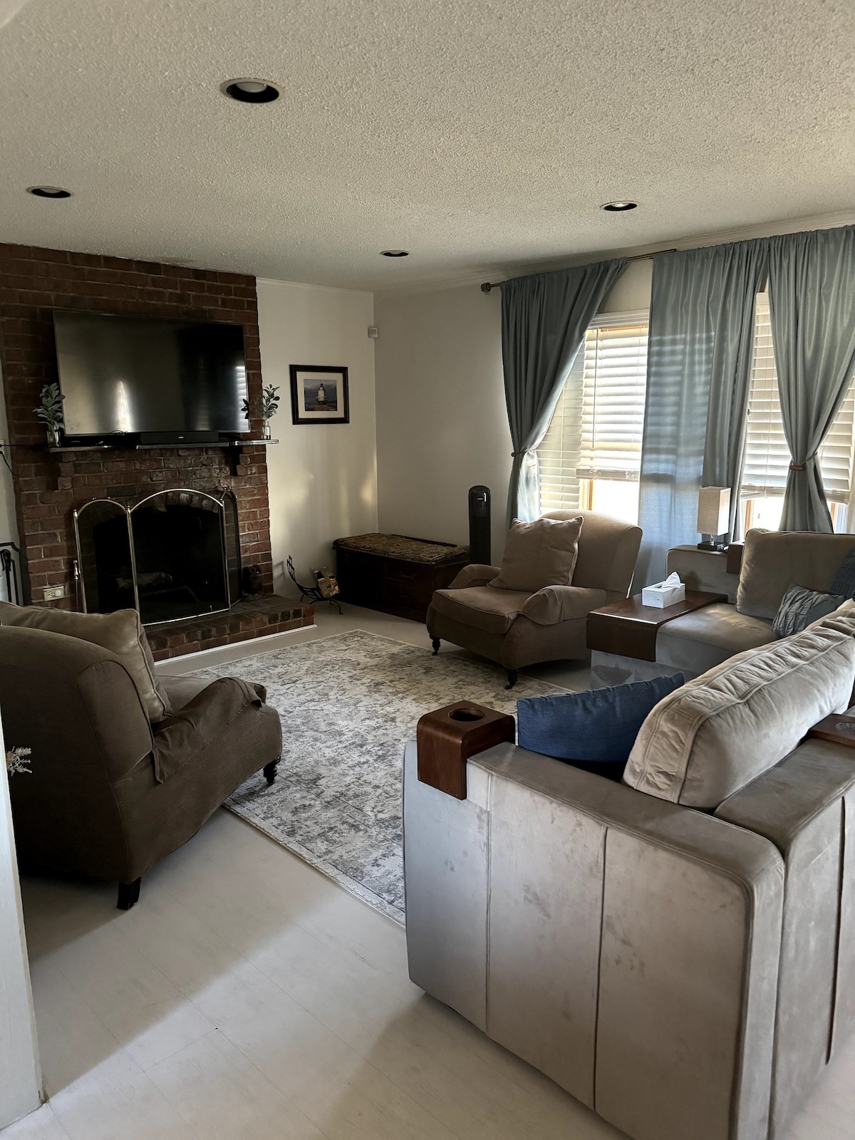A cozy living room features soft seating arrangements, including two plush armchairs and a couch, arranged around a central area rug. A fireplace with a mounted television above serves as a focal point, while natural light filters through the large window adorned with blue curtains.