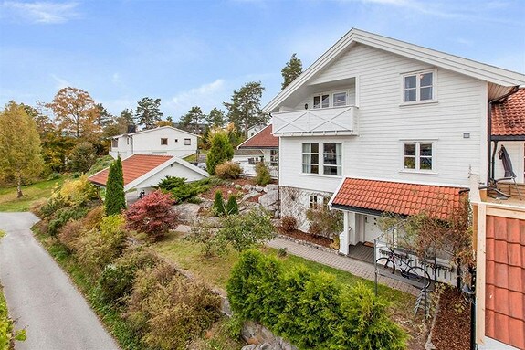 The exterior of a charming white house is presented, featuring a sloped red-tiled roof and a balcony off the upper level. Surrounding the home are well-maintained gardens with various shrubs and trees, contributing to a serene environment and inviting entrance.