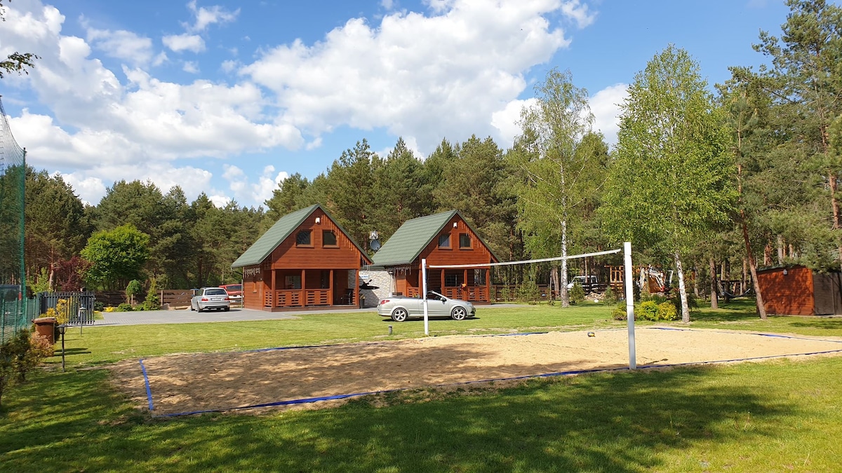 Two wooden cabins with green roofs are set within a grassy area, surrounded by trees. A sand volleyball court is visible in the foreground, lined with a blue boundary. A parking area for cars is located nearby, contributing to the outdoor space.