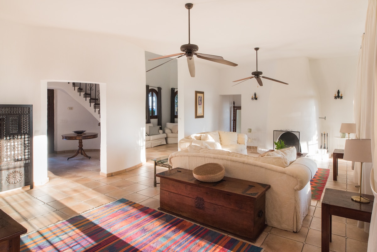 A spacious living area features a light, airy design, with a large white sofa surrounded by natural light from windows. A colorful woven rug adds a touch of warmth to the tiled floor. Two ceiling fans and a log-burning fireplace enhance the comfort of the space.