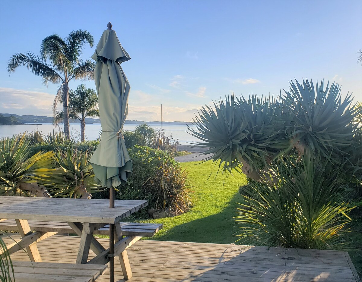 A wooden deck features a picnic-style table with an upright umbrella, facing a tranquil view of the sea. Lush greenery, including palm trees and coastal plants, surrounds the area, enhancing the peaceful outdoor setting with a glimpse of the beach in the distance.