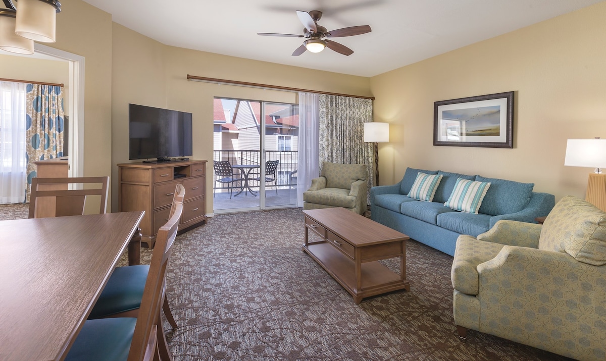 A welcoming living area is featured, furnished with a teal couch and a light-toned armchair. A wooden coffee table sits in front, and a flat-screen television is mounted on the wall. Large sliding doors lead to a patio area, inviting natural light into the space.