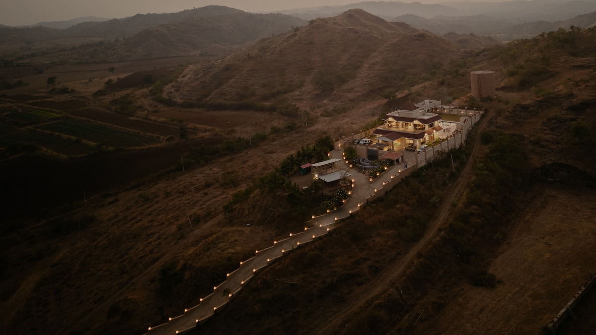 An aerial view captures a luxurious villa nestled amidst rolling hills. A winding pathway lined with lights leads to the property, which is surrounded by rustic landscapes and scattered greenery, showcasing a serene and remote setting.
